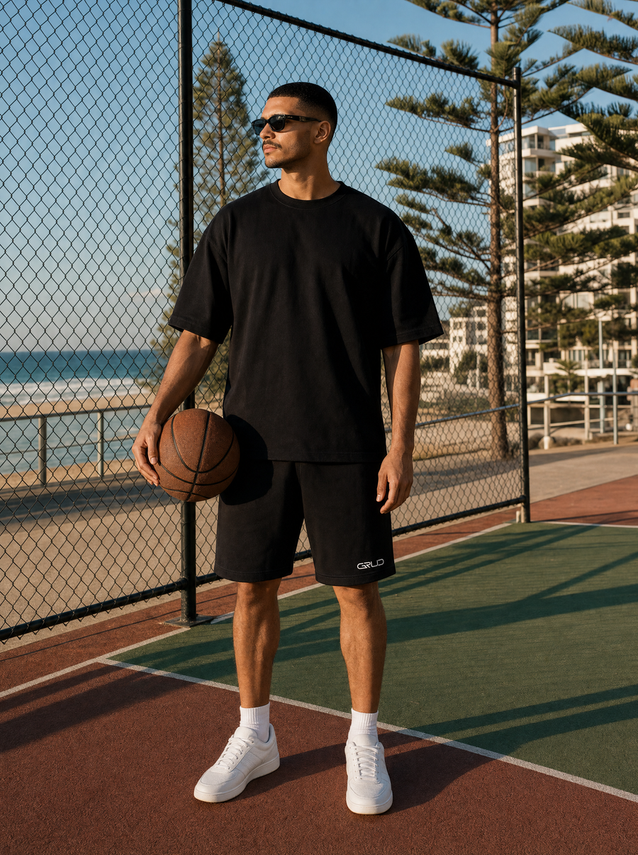 Man in the GRUD tee and shorts at an outdoor basketball court near the beach